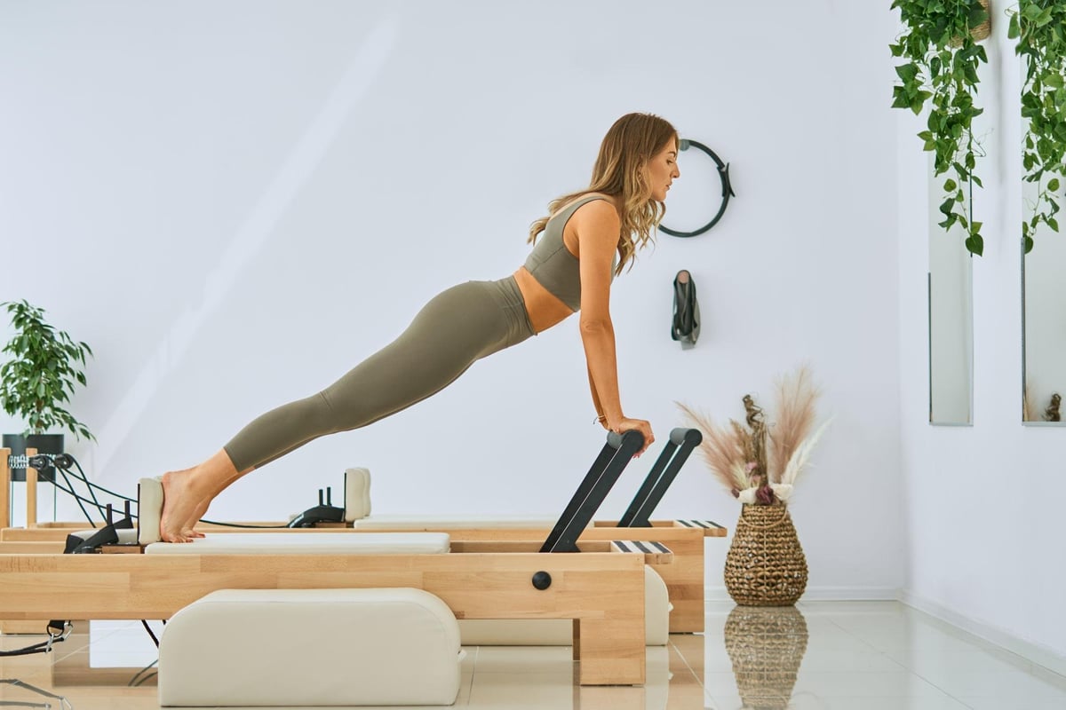Woman in athletic wear performing a plank exercise on a wooden Pilates reformer in a bright studio with plants