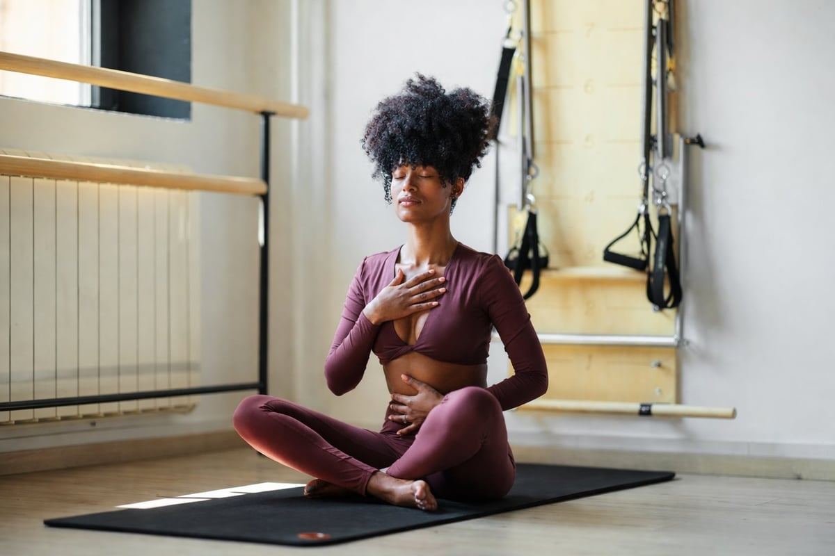 Woman in purple workout outfit sitting on yoga mat in gym, hand on chest, with exercise equipment visible in background